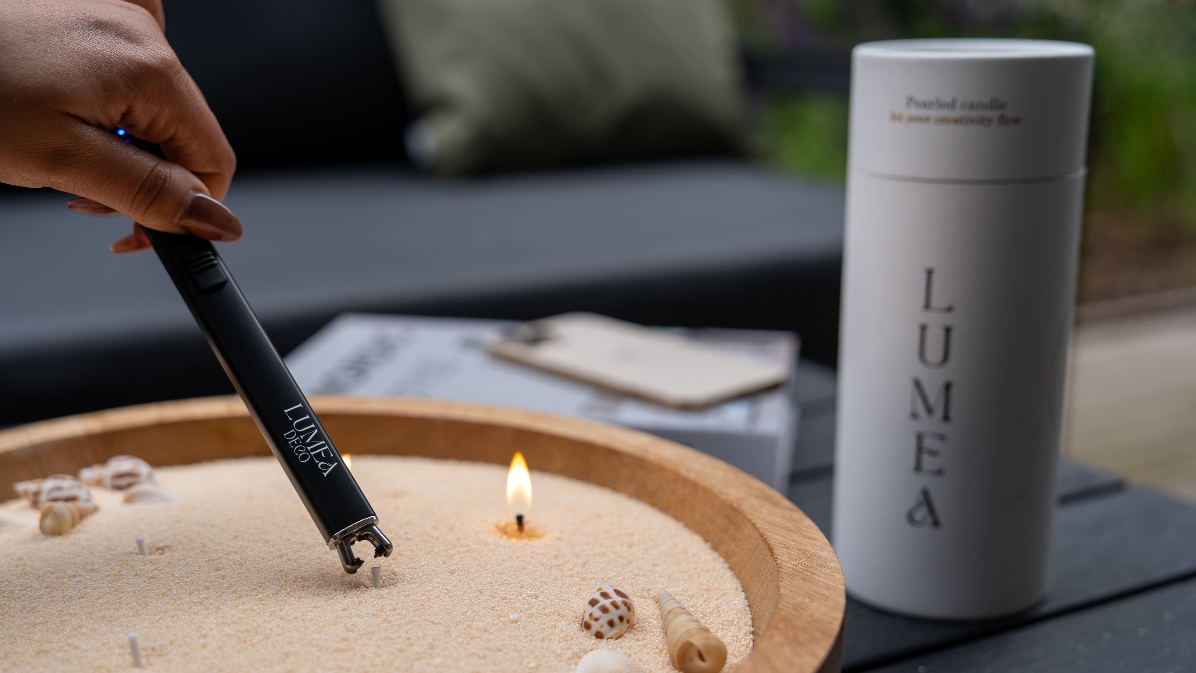 Bougie perlée Lumea allumée avec un briquet USB dans un contenant décoratif rempli de cire perlée et de coquillages, illustrant la personnalisation et la sécurité du concept.