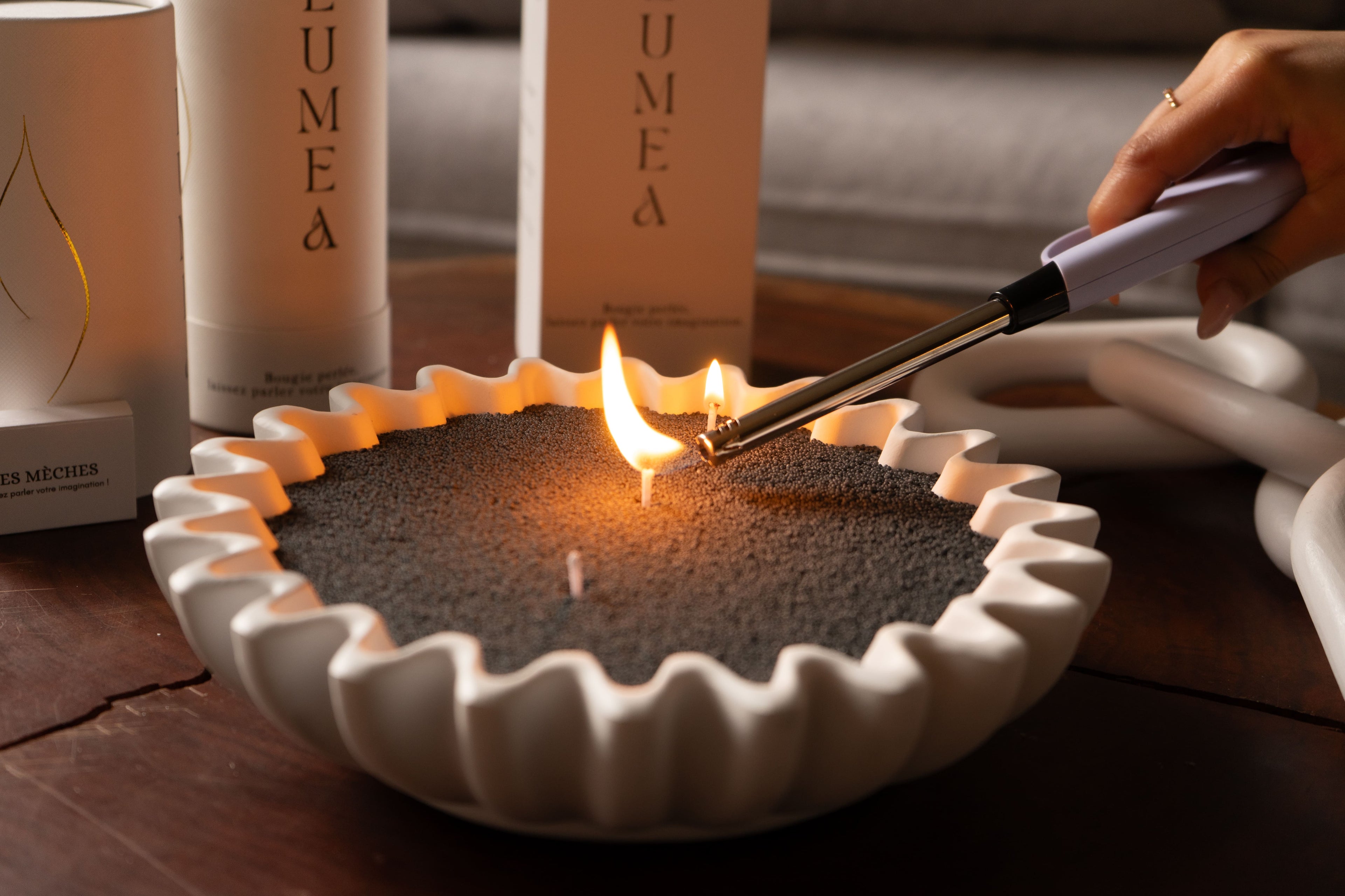 Allumage d’une bougie perlée noire Lumea dans un récipient décoratif blanc, avec packaging visible en arrière-plan – concept de bougie rechargeable, design et personnalisable.