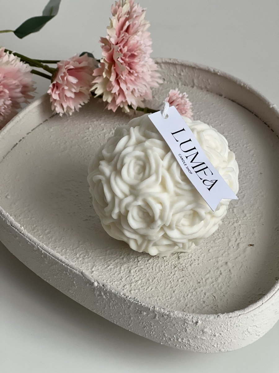 Bougie décorative en forme de boule composée de roses blanches sculptées, posée sur un plateau texturé beige avec fleurs roses en arrière-plan, création Lumea Deco.