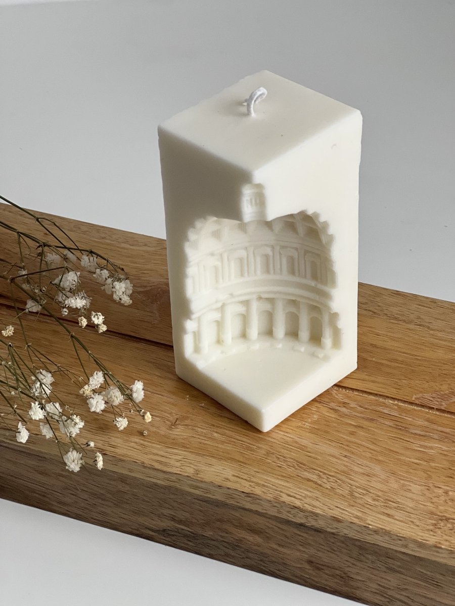 Bougie blanche en forme hexagonale avec relief du Colisée romain, posée sur une planche en bois avec fleurs séchées, ambiance artisanale et élégante, collection Lumea Deco.