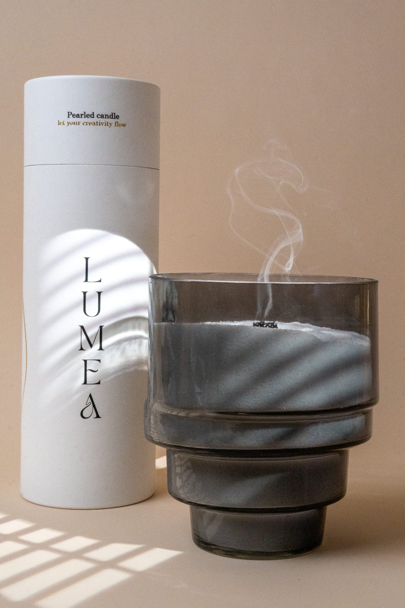Bougie perlée blanche dans un récipient cylindrique en verre fumé à étages, fumée douce s’échappant, posée à côté du tube signature Lumea Deco.