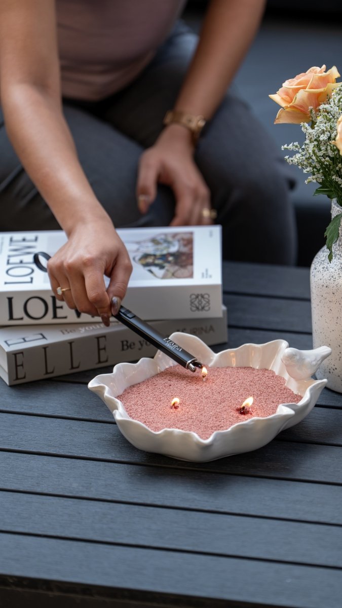 Bougie perlée rose dans un récipient en forme de coquillage blanc, allumée sur une table extérieure avec des livres et un bouquet ; idéale pour une ambiance élégante ou comme idée cadeau originale.