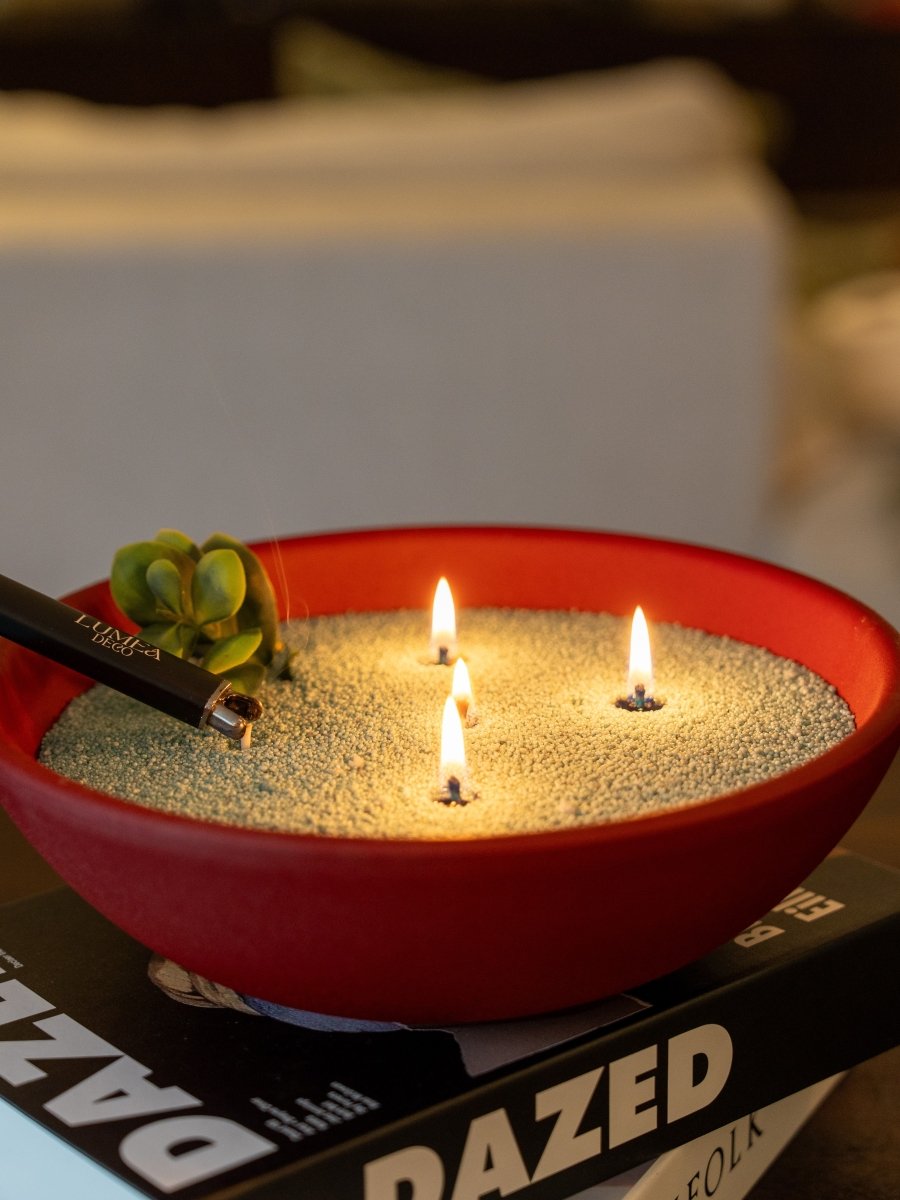 Bougie perlée allumée dans un bol rouge rempli de cire perlée  avec trois flammes, posée sur des livres déco, briquet USB Lumea à côté – ambiance chaleureuse et moderne.