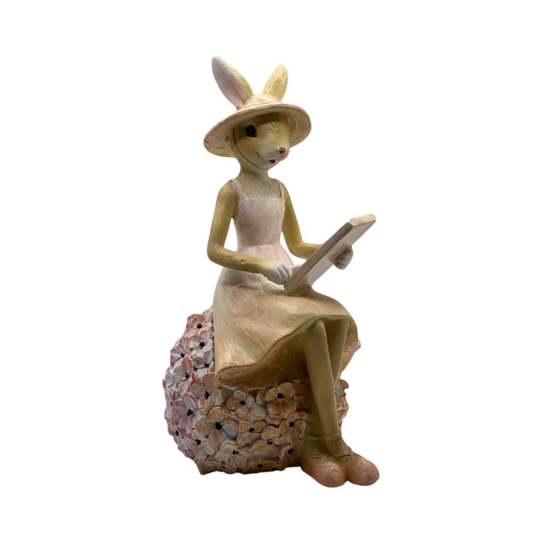 Figurine lapin de Pâques en résine, représentant une femme assise avec chapeau et livre. Finition bois sculpté, base fleurie. Décoration originale pour printemps ou idée cadeau.