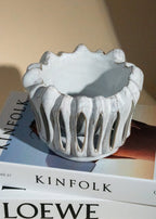 Photophore en céramique blanche à structure ajourée et forme organique, posé sur des livres Kinfolk et Loewe dans une ambiance lumineuse naturelle.