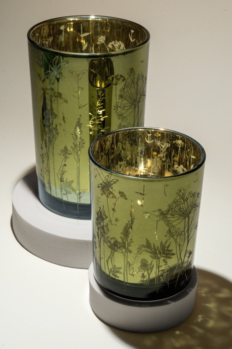 Duo de photophores en verre vert translucide, ornés de motifs floraux délicats, avec intérieur doré réfléchissant, posés sur socles ronds blancs. objet déco