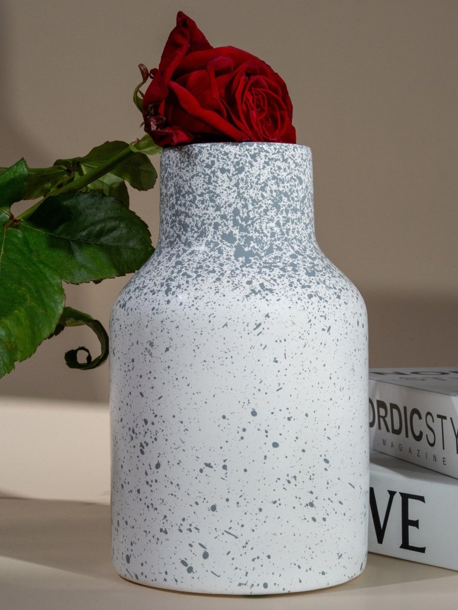 Vase en céramique blanc tacheté de gris avec rose rouge insérée, posé sur une table décorative avec magazines, style nordique et moderne.