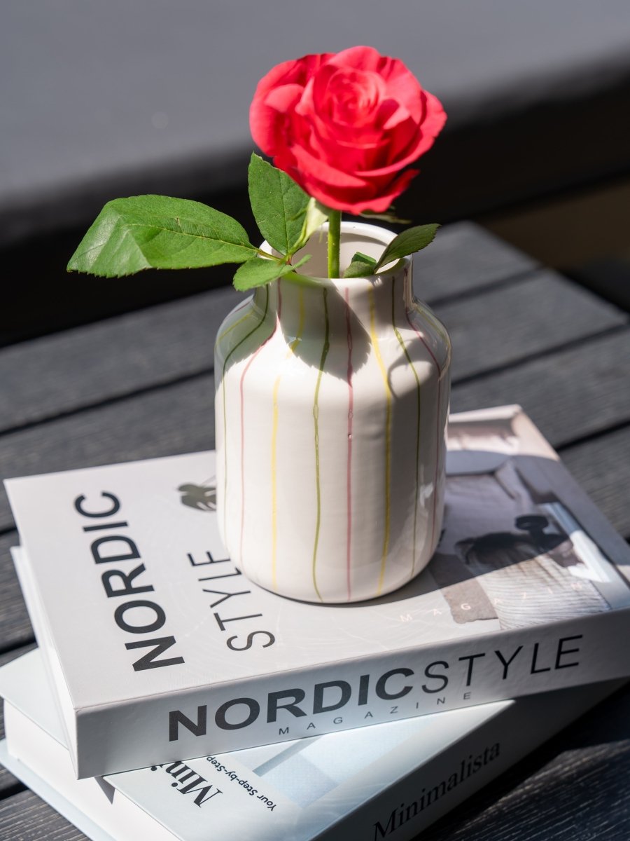 Petit vase en céramique blanche à rayures verticales jaunes, vertes et roses, contenant une rose rouge, posé sur deux magazines "Nordic Style" en extérieur.