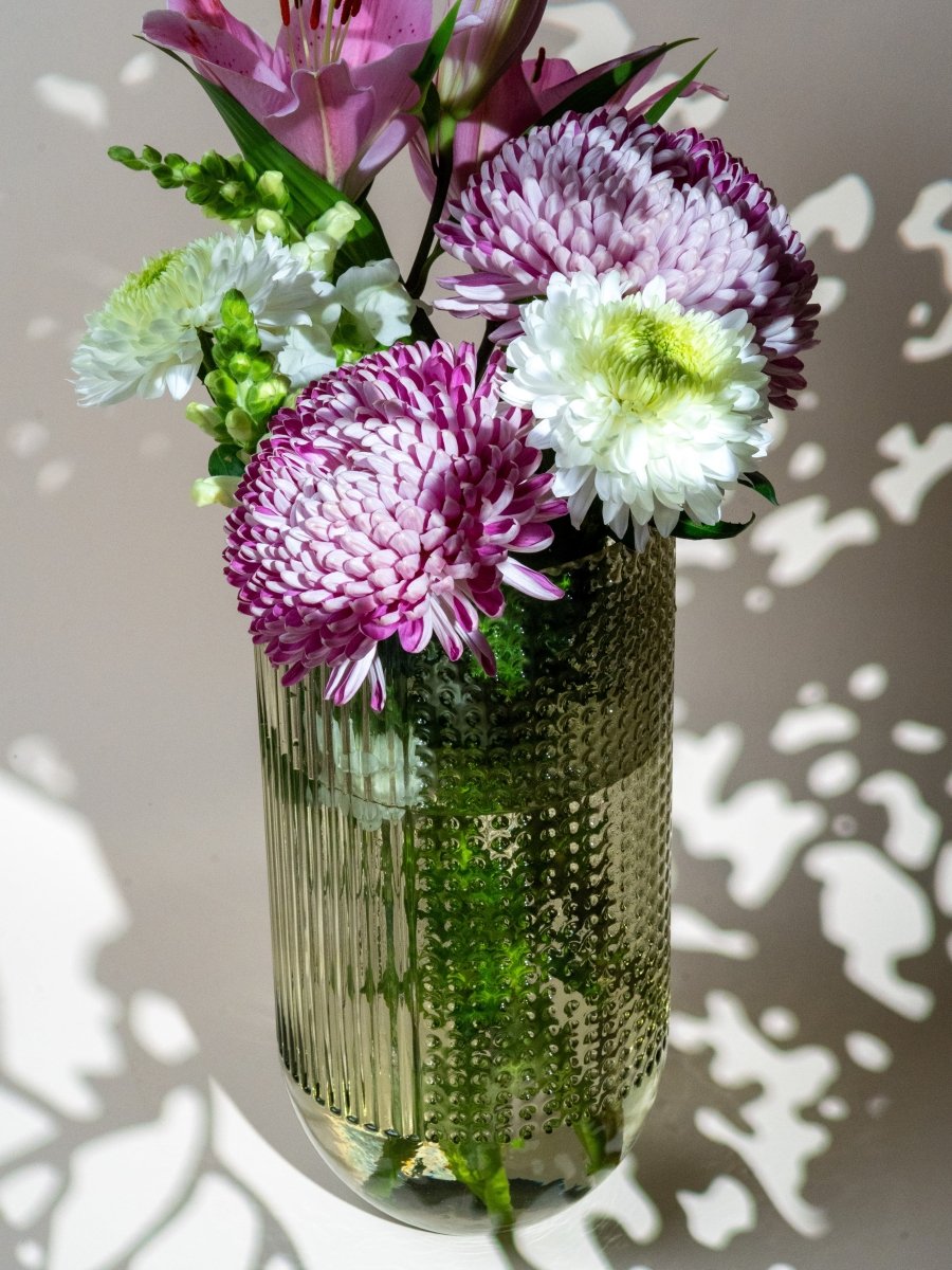 Vase en verre transparent à reliefs striés et en pois, contenant un bouquet de fleurs fraîches aux tons violets, blancs et roses – décoration florale raffinée, parfaite à offrir.