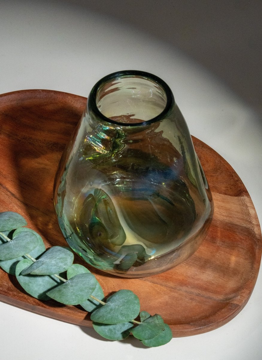 Vase en verre soufflé transparent aux reflets verts, forme organique irrégulière, posé sur plateau en bois avec feuillage – objet déco nature et artisanal, parfait pour fleurs.