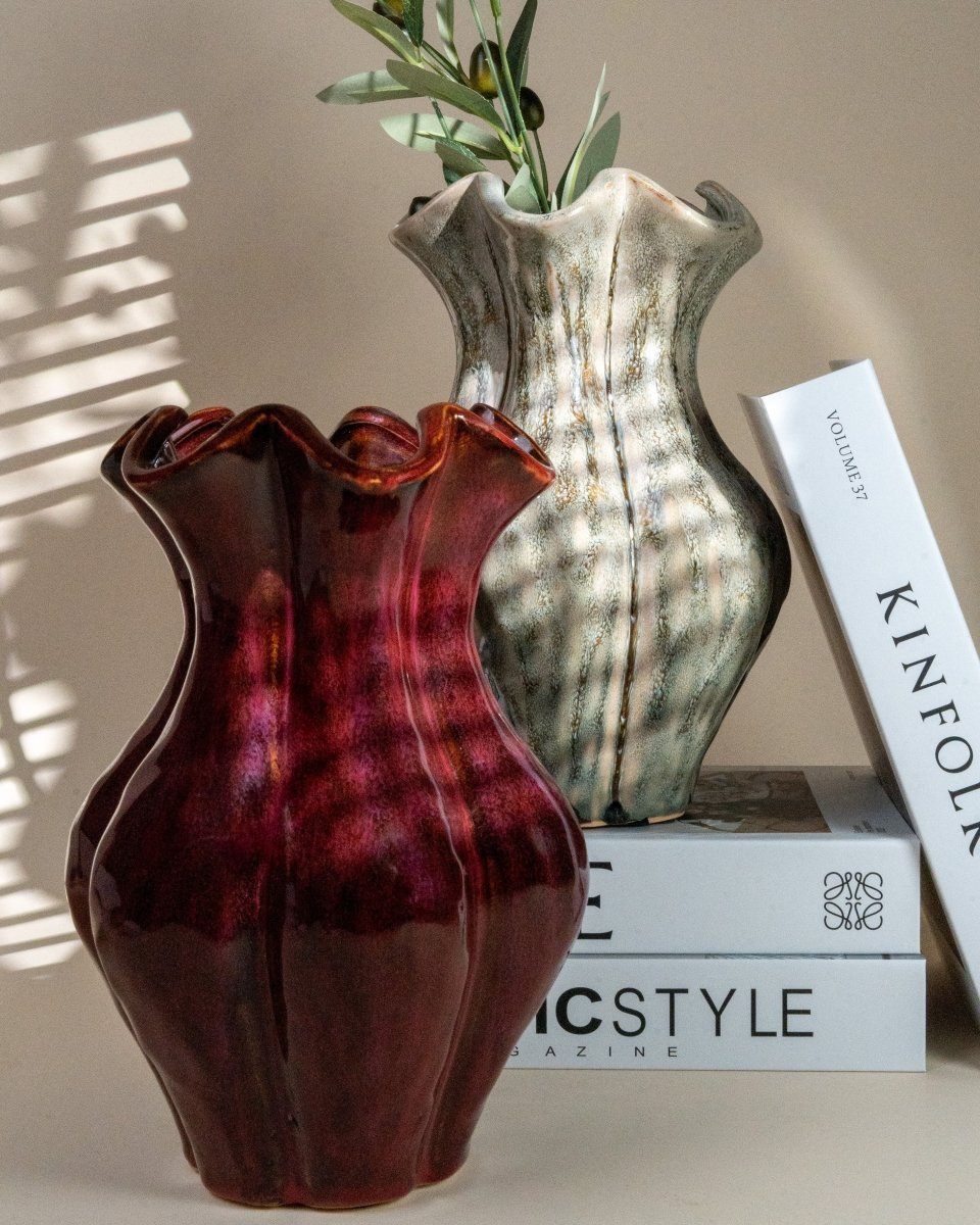 Deux vases en céramique à col ondulé, l’un rouge foncé et l’autre vert moucheté, posés sur des livres de décoration dans un intérieur lumineux ; parfaits pour accueillir des fleurs ou offrir en cadeau déco.